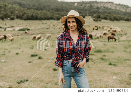 A woman in a plaid shirt and hat stands in a field with a herd of sheep 126490884