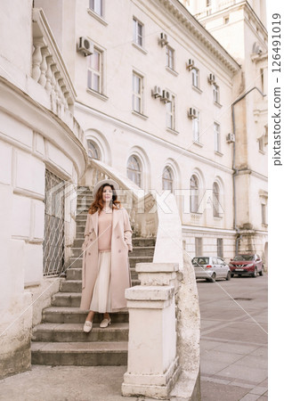 woman in elegant coat and hat against an intricate architectural backdrop, harmoniously blending modern fashion with historical allure. The soft daylight adds to its timeless appeal. 126491019