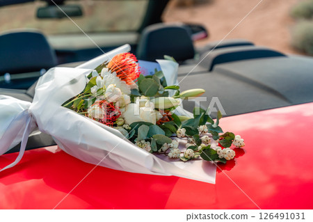 A bouquet of flowers is sitting on the hood of a red car. The flowers are white and red, and they are arranged in a vase. Concept of romance and love. A bouquet of flowers is sitting on the hood of a red car. The flowers are white and red, and they are arranged in a vase. Concept of romance and love. 126491031