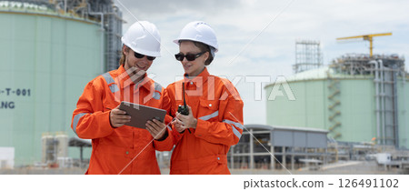 Professional engineer wearing orange suit and white hard hat stands using tablet in at liquefied natural gas facility industrial structures petroleum. Survey planning for gas transmission projects Professional engineer wearing orange suit and white hard hat stands using tablet in at liquefied natural gas facility industrial structures petroleum. Survey planning for gas transmission projects 126491102