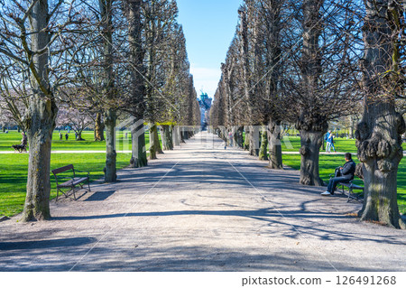 Visitors stroll through the lined pathways of Rosenborg Castle Gardens in Copenhagen. Trees cast shadows on the gravel as guests enjoy the peaceful spring atmosphere. 126491268