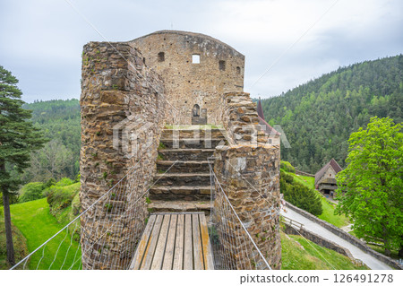 Visitors walk across the bridge of Velhartice Castle, surrounded by lush greenery and steep hills. The ancient stone structure offers a glimpse into history and stunning views. 126491278