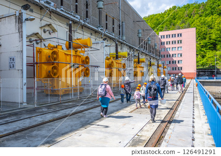 Tourists walk along the pathway at Orlik Dam in Czechia, observing the hydroelectric power station's generators and surrounding nature on a sunny day. Tourists walk along the pathway at Orlik Dam in Czechia, observing the hydroelectric power station's generators and surrounding nature on a sunny day. 126491291