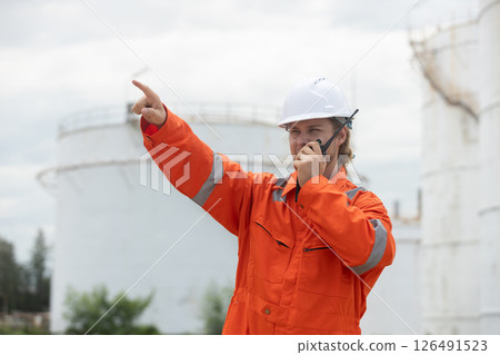 worker in orange safety suit communicates via radio, directing activities at an industrial site with storage tanks. worker in orange safety suit communicates via radio, directing activities at an industrial site with storage tanks. 126491523