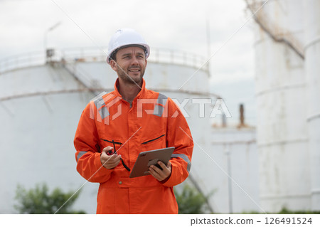 Worker in safety gear using tablet near industrial storage oil tanks refining petrochemical industry factory. Worker in safety gear using tablet near industrial storage oil tanks refining petrochemical industry factory. 126491524
