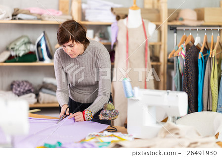 Female dressmaker cutting fabric with scissors while working in sewing workshop 126491930