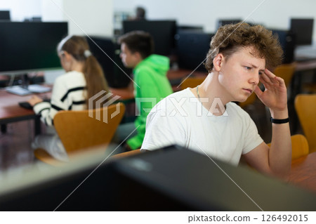 Tired schoolboy with a sore head sits at a computer in classroom Tired schoolboy with a sore head sits at a computer in classroom 126492015