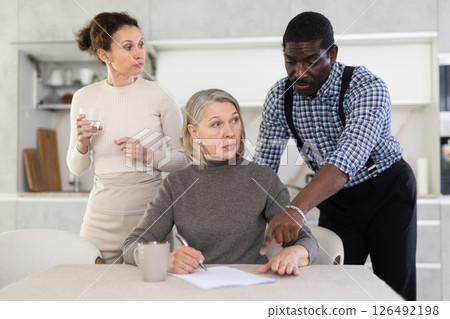 Old woman compiling testament while middle-aged man and woman competing with each other in the kitchen 126492198