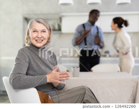 Smiling old woman sitting at the kitchen table with her back to couple quarreling together 126492260