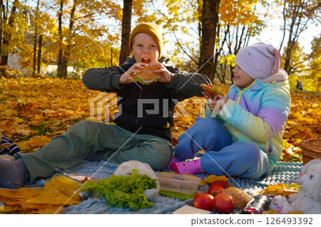 A boy and a girl sit closely on a soft plaid, enjoying homemade treats as autumn leaves dance around them in the crisp afternoon air. 126493392
