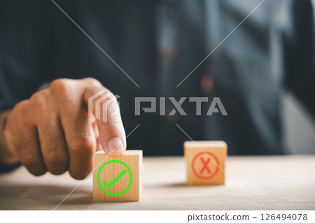 Finger of businessman touches wooden cubes with check marks showing right and wrong choices. Communicates choice validation in decision-making. Think With Yes Or No Choice. 126494078