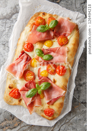 Pinsa Romana with cherry tomatoes, ham, mozzarella cheese and basil closeup on parchment paper. Vertical top view 126494218