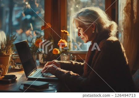 Senior woman typing on laptop at home with warm sunlight and flowers creating a cozy atmosphere Senior woman typing on laptop at home with warm sunlight and flowers creating a cozy atmosphere 126494713