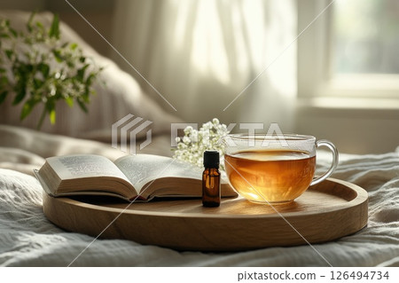 Warm cup of herbal tea, open book, and essential oil bottle on wooden tray in cozy setting 126494734