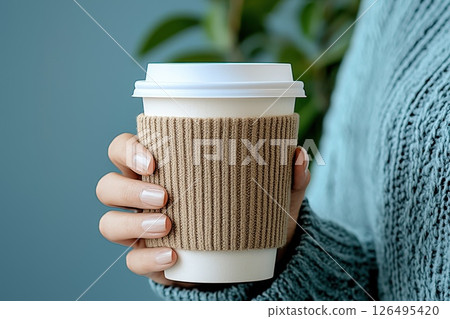 Cozy Hand Holding Coffee Cup with Sleeve in Warm Knit Sweater Against Soft Background Cozy Hand Holding Coffee Cup with Sleeve in Warm Knit Sweater Against Soft Background 126495420