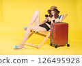 Cheerful tourist girl sitting with suitcase and smartphone on vacation, enjoying holiday travel vibes on yellow backdrop in swimwear and casual beach outfit 126495928