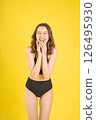 Stylish young woman in black bikini smiling and laughing on yellow background, posing playfully for summer travel, swimwear, or vacation-themed concepts 126495930