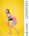 Playful young woman in black swimsuit holding colorful float and eating ice cream cone, posing happily for summer vacation studio shot on bright yellow background 126495932