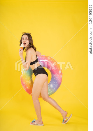 Playful young woman in black swimsuit holding colorful float and eating ice cream cone, posing happily for summer vacation studio shot on bright yellow background 126495932