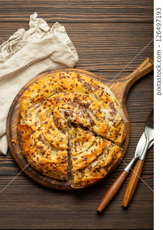 Burek with cut piece made of filo dough with filling on cutting board, dark brown wooden rustic background top view. Traditional savoury spiral pie of Balkans, Middle East and Central Asia Burek with cut piece made of filo dough with filling on cutting board, dark brown wooden rustic background top view. Traditional savoury spiral pie of Balkans, Middle East and Central Asia 126497193
