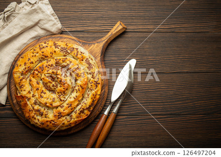 Burek made of filo dough with filling on cutting board, dark brown wooden rustic background top view. Traditional savoury spiral pie of Balkans, Middle East and Central Asia. Space for text Burek made of filo dough with filling on cutting board, dark brown wooden rustic background top view. Traditional savoury spiral pie of Balkans, Middle East and Central Asia. Space for text 126497204
