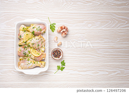 Raw uncooked chicken legs with seasonings, herbs, lemon in white ceramic casserole top view on light wooden rustic background. Preparing healthy meal with marinated chicken drumsticks. Space for text 126497206