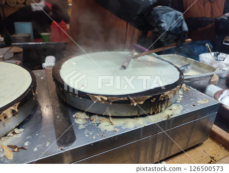 Frying pancakes. Person chef spreading liquid batter on round frying pan spatula. Cooking food on street. Festivities festival. Street food fried batter dough. 126500573
