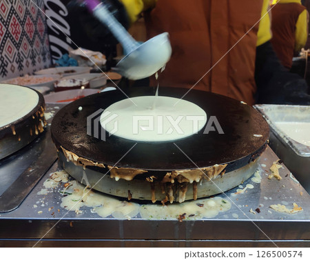 Person pouring ladle of batter into round pan for frying pancakes. Cook making pancakes from fried dough. Frying pancakes outdoors. Cooking street food at festival. Cooking meals on street 126500574