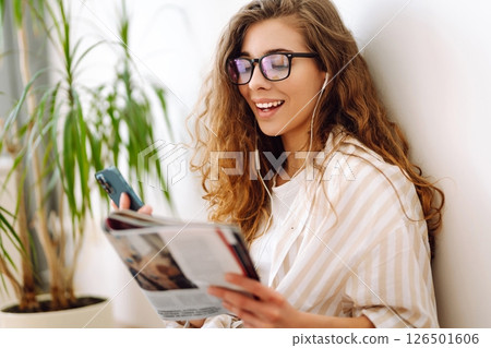 Attractive woman sits on floor in apartment with book on headphones while being distracted by phone. 126501606