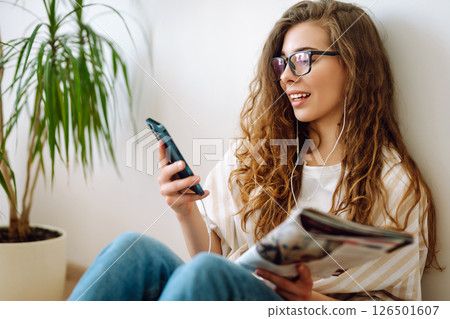Attractive woman sits on floor in apartment with book on headphones while being distracted by phone. 126501607