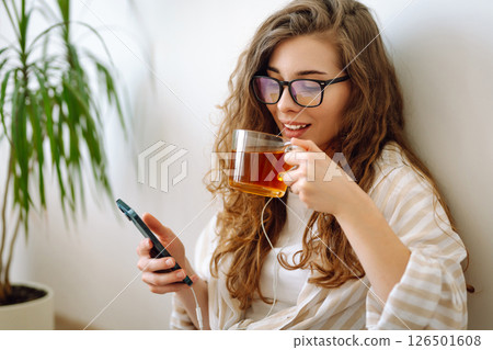 A young curly woman reads a book and drinks coffee or tea while sitting on floor in the living room. 126501608