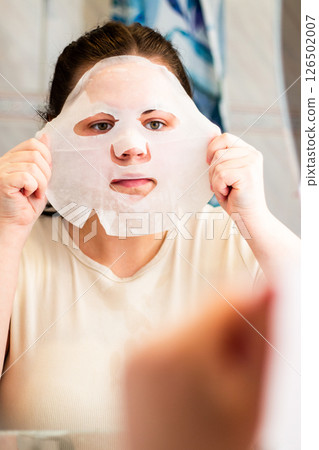 Close up of female adjusting sheet mask on face in modern bathroom. Ideal for cosmetic ads or spa treatment visuals. Close up of female adjusting sheet mask on face in modern bathroom. Ideal for cosmetic ads or spa treatment visuals. 126502007