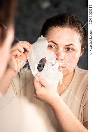 Woman removing sheet face mask in bathroom mirror, revealing fresh glowing skin. Perfect for after care skincare routines and beauty product promotions. 126502011