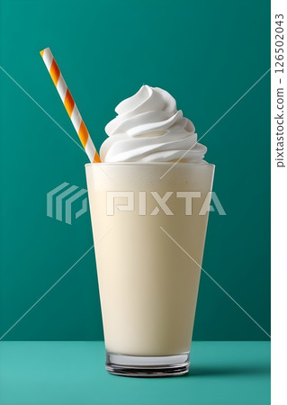 Classic vanilla milkshake in a tall glass against a vibrant turquoise background. The beverage features a generous swirl of whipped cream on top and is served with an orange. 126502043
