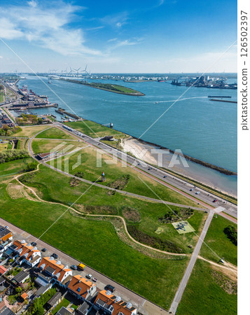 Aerial view of a coastal town with green parks, residential areas, a riverfront road, and busy shipping lanes. Wind turbines and industrial port facilities are visible across the water. 126502397