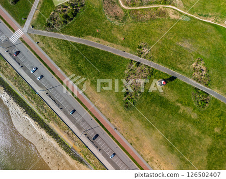 Aerial view of a coastal road with parked cars, a footpath through green grassland, and a small red lighthouse structure in a peaceful natural setting. Aerial view of a coastal road with parked cars, a footpath through green grassland, and a small red lighthouse structure in a peaceful natural setting. 126502407