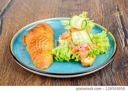 On half of a croissant are ingredients for filling of lettuce leaves, pieces of red fish and cucumbers. 126503058