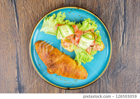Top view of croissants with filling ingredients of lettuce leaves, pieces of red fish and cucumbers. 126503059