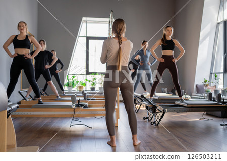 Young caucasian women exercising on pilates reformers in a bright studio. Their workout focuses on core strength and flexibility in a modern fitness environment. Young caucasian women exercising on pilates reformers in a bright studio. Their workout focuses on core strength and flexibility in a modern fitness environment. 126503211