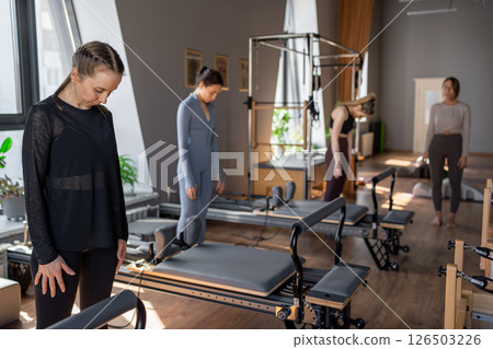 Young caucasian woman doing roll down exercise in a pilates bright studio. Their workout focuses on core strength and flexibility in a modern fitness environment. 126503226