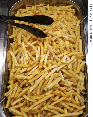 Golden french fries filling metal serving tray, crispy surface gleaming, black tongs resting alongside golden potatoes 126503314