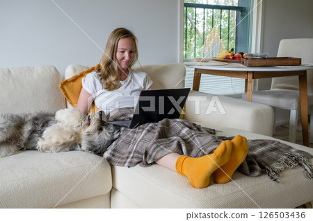 Woman with bright socks sitting on cream sofa with blanket and dog and using laptop Woman with bright socks sitting on cream sofa with blanket and dog and using laptop 126503436