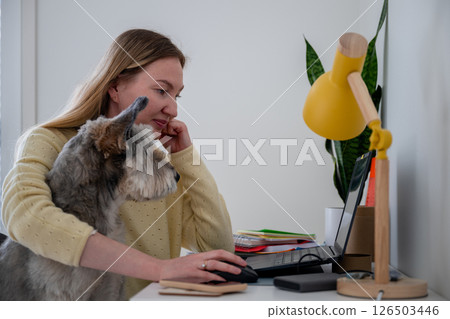 Woman works on laptop with dog on lap in home office, concept of remote work and pet companionship 126503446
