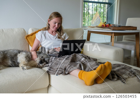 Woman with bright socks sitting on cream sofa with blanket and dog and using laptop Woman with bright socks sitting on cream sofa with blanket and dog and using laptop 126503455