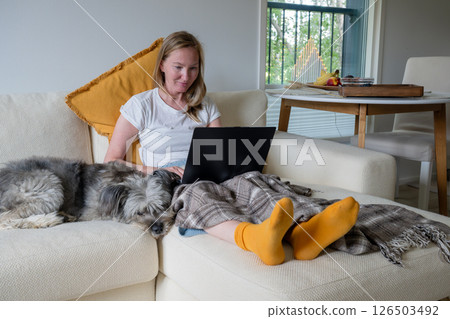 Woman with bright socks sitting on cream sofa with blanket and dog and using laptop 126503492