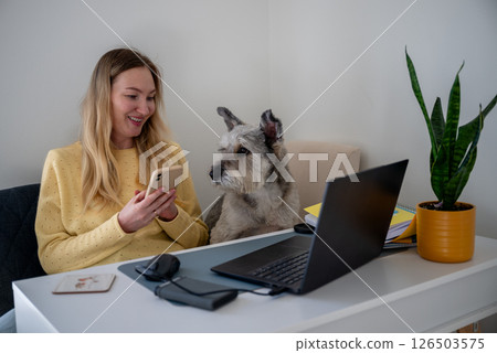 Woman in yellow sweater using phone with dog beside her at home office desk, concept of remote work Woman in yellow sweater using phone with dog beside her at home office desk, concept of remote work 126503575