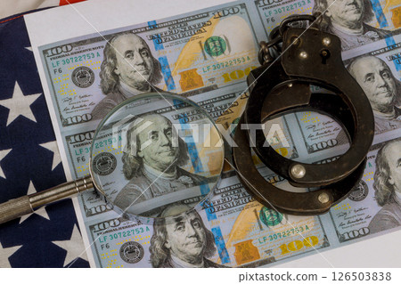 Handcuffs rest top multiple US hundred dollar bills while magnifying glass focuses on one bill serious mood evokes an investigative atmosphere. 126503838