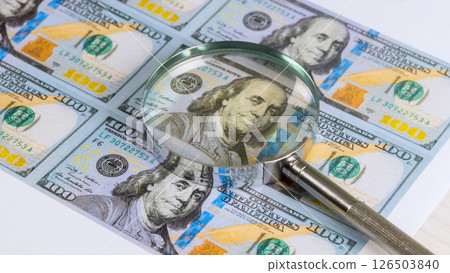 Magnifying glass highlights details of US hundred dollar bill featuring Benjamin Franklin creates analytical investigative mood. Magnifying glass highlights details of US hundred dollar bill featuring Benjamin Franklin creates analytical investigative mood. 126503840