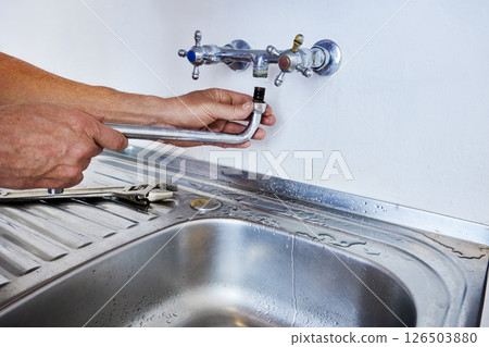 Person is installing faucet spout into wall-mounted tap above kitchen sink while using hands and water is visible on stainless steel surface. Person is installing faucet spout into wall-mounted tap above kitchen sink while using hands and water is visible on stainless steel surface. 126503880