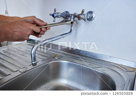 Person is tightening faucet spout to wall-mounted tap using adjustable wrench while standing over kitchen sink with visible metal drainer. Person is tightening faucet spout to wall-mounted tap using adjustable wrench while standing over kitchen sink with visible metal drainer. 126503889